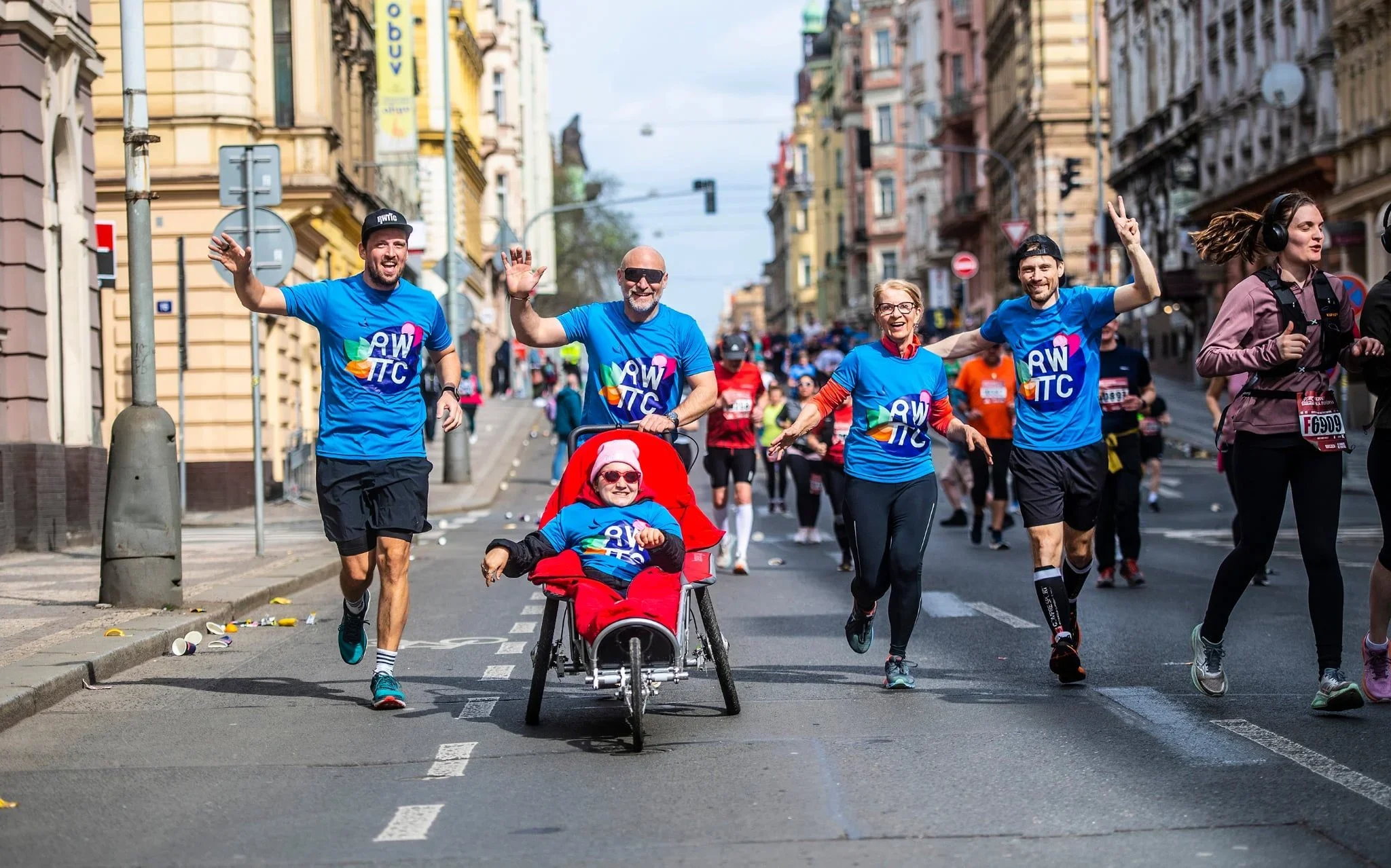 Běžci RWTTC mávají a tlačí dítě v červeném vozíku ulicemi Prahy